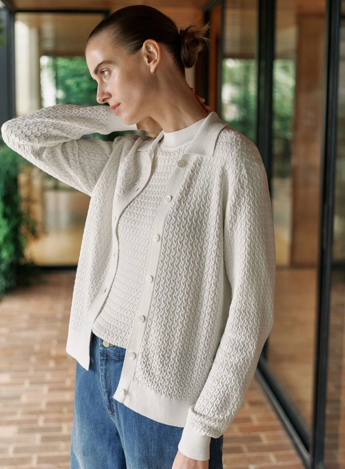 A model with her hair in a sleek bun wearing the Trenery Micro-cable knit set in white. The outfit features a textured, short-sleeved crew neck top layered under a matching long-sleeved cardigan with a collar and white buttons. It is styled casually with blue denim jeans. Best knitwear brands Australia article.