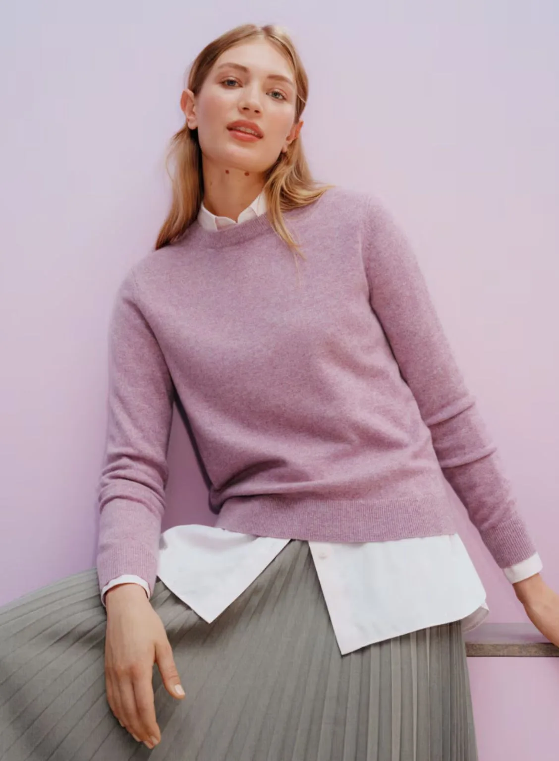 A model with blonde hair pulled back, wearing a Uniqlo cashmere crew neck sweater in a soft lilac purple. The jumper is layered over a white collared shirt and paired with a dark grey pleated midi skirt. She is standing outdoors against a minimalist concrete and glass building. Best knitwear brands Australia article.