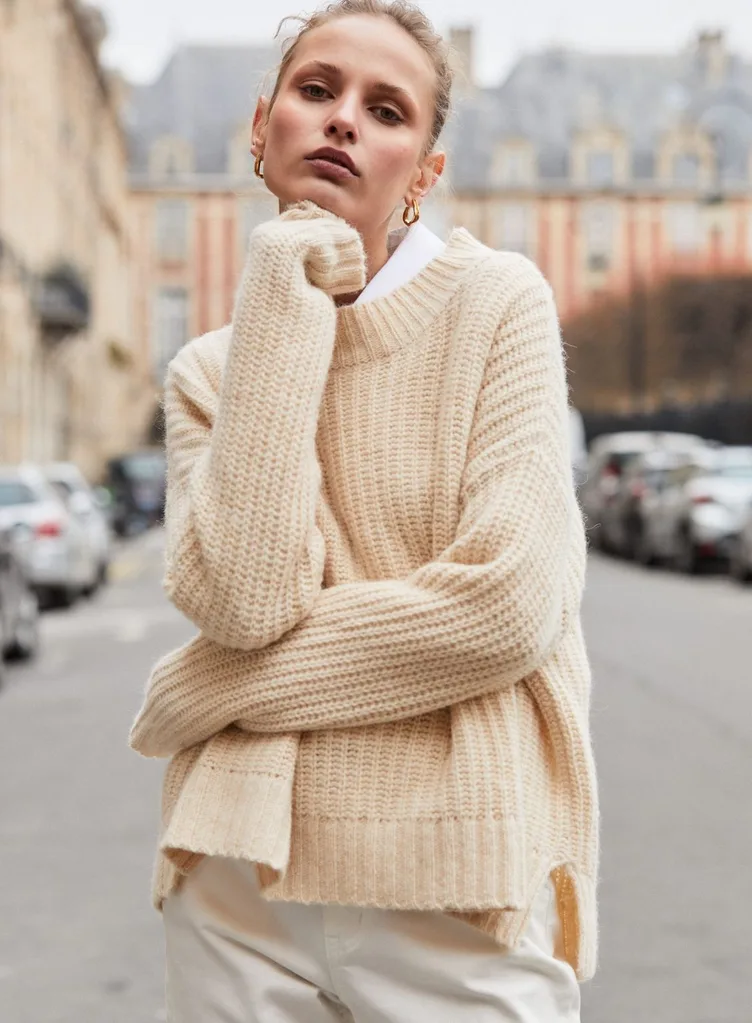 A model standing on a city street wearing the Viktoria & Woods Nevermore Knit in a pale "butter" yellow hue. The chunky, ribbed crew neck jumper features an oversized fit with side splits and is layered over a white collared shirt and cream trousers. Best knitwear brands Australia article.