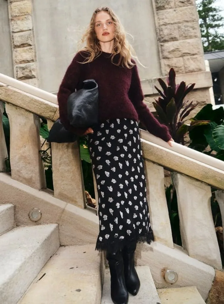 A model with long wavy blonde hair standing on stone steps, wearing a fuzzy, deep burgundy crew neck jumper from Witchery. The outfit is paired with a black floral midi skirt featuring a lace hem, black leather boots and a large black slouchy leather bag. Best knitwear brands Australia article.
