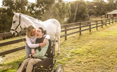 Woman hugging a man in a wheelchair, with a white horse standing beside them on a sunny day in a grassy field.