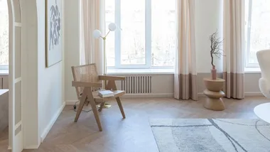 A minimalist living room with a wooden chair, side table, tall lamp, abstract painting, and light curtains over large windows.