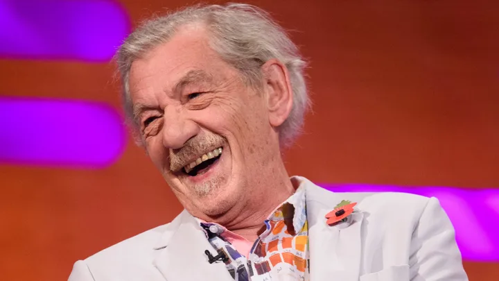 A man in a light-colored jacket laughs, wearing a colorful shirt and poppy pin, with a vibrant purple background.