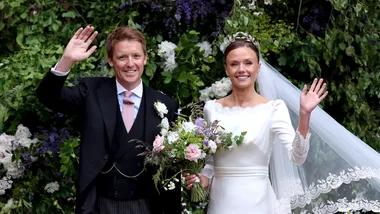 Bride and groom smiling and waving at their wedding, surrounded by a lush floral backdrop.
