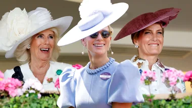 Three women smiling, dressed elegantly in pastel and floral outfits with decorative hats, standing amidst pink flowers.