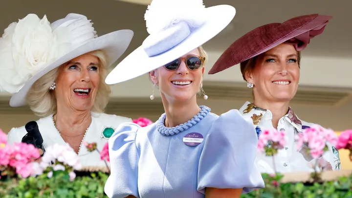 Three women smiling, dressed elegantly in pastel and floral outfits with decorative hats, standing amidst pink flowers.