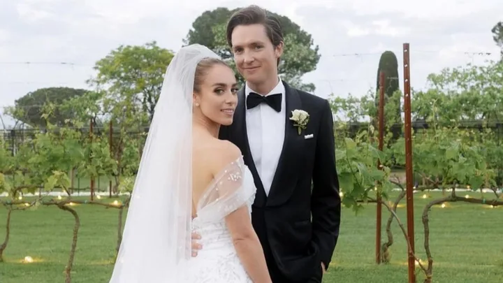 Bride in white gown and veil, groom in black tuxedo, posing in a vineyard setting.