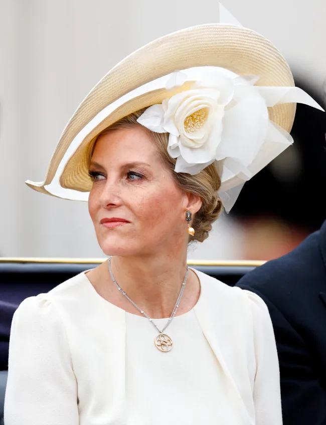 A woman in a white dress and elegant hat with a large flower, sitting and looking to the side.