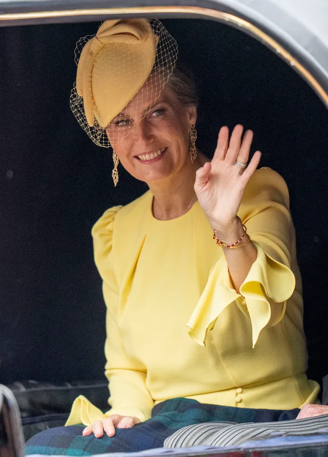 Woman in a yellow outfit with matching hat and veil, seated in a vehicle, smiles and waves.