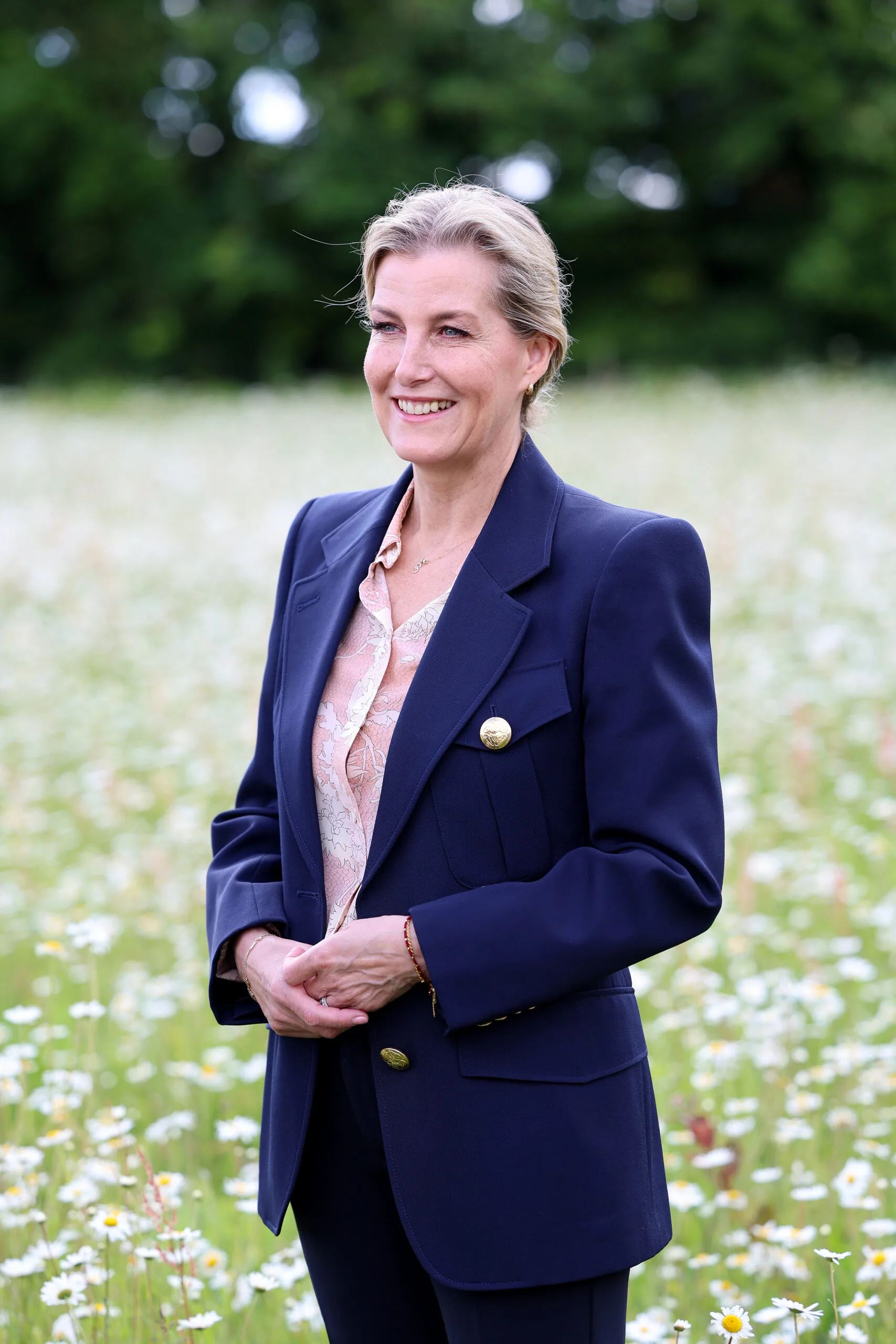 Elegant woman in navy blazer and floral blouse stands smiling in a field of white flowers.