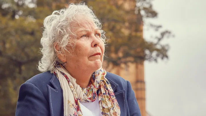 Elderly woman with curly white hair and scarf looks contemplatively upwards, blurry trees and building in background.