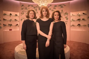 Three women in black dresses stand in a chic shoe store with floral wallpaper and a crystal chandelier overhead.