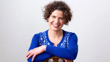 Smiling woman with curly hair in a blue patterned sweater, leaning on a chair in front of a light background.