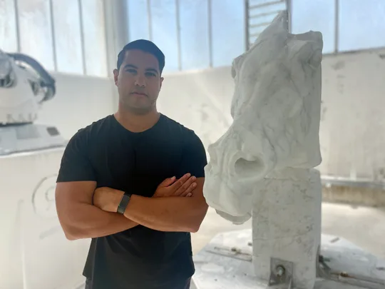 Man standing beside large marble horse head sculpture in a bright studio.