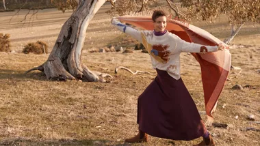 Person joyfully runs with a patterned blanket in a grassy field beside a twisted tree.
