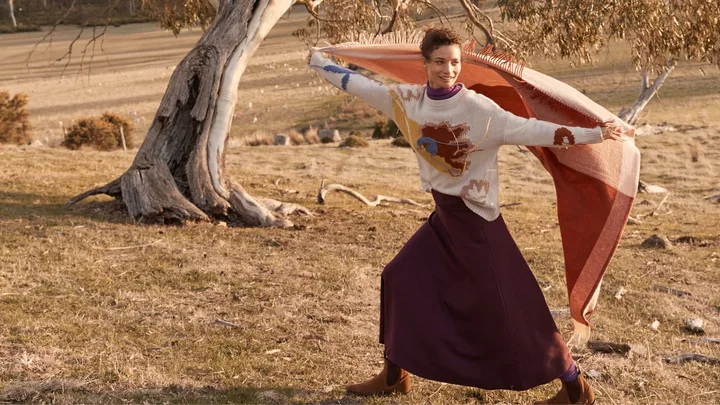 Person joyfully runs with a patterned blanket in a grassy field beside a twisted tree.