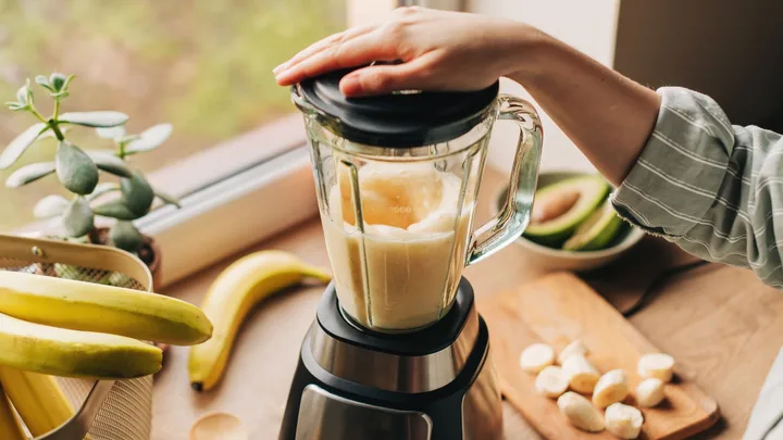Person blending bananas in a kitchen, with avocados and bananas on a wooden countertop.