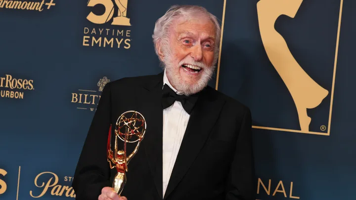 Elderly man in a tuxedo holds a Daytime Emmy Award, smiling, with a blue and gold Emmy backdrop.