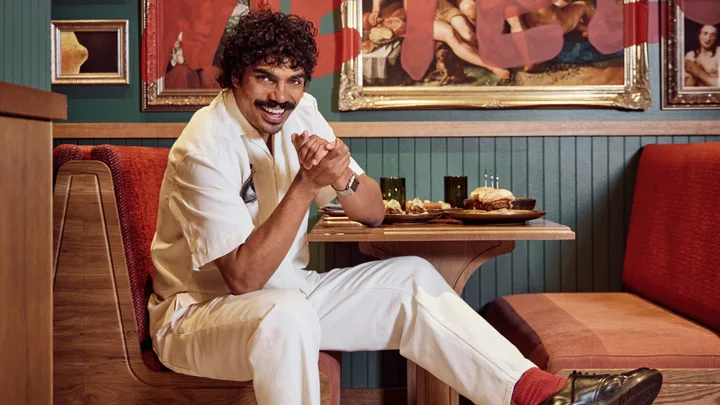 Man in white outfit sitting at a restaurant booth, smiling, with framed art and food on the table.