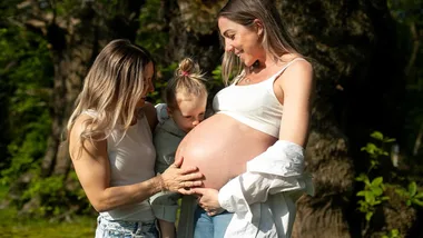 Two women and a child outside; one woman pregnant, the child touching her belly, all smiling warmly.
