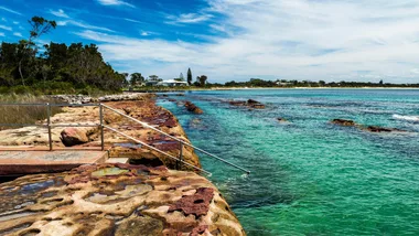 Rocky seaside with clear turquoise water and distant shoreline with trees and houses.