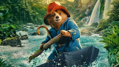 A bear in a blue coat and red hat paddles a makeshift raft on a river in a lush, green jungle setting.