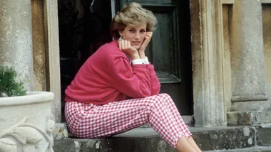 A woman in a pink sweater and checkered pants sits on stone steps, resting her chin on her hands.