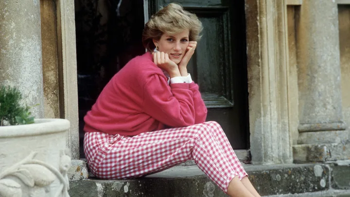 A woman in a pink sweater and checkered pants sits on stone steps, resting her chin on her hands.