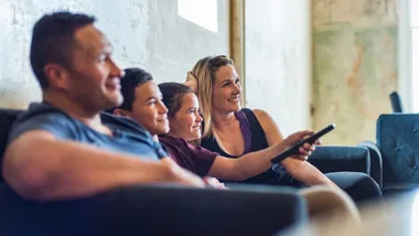 A family of four sits on a couch, smiling and watching TV, with the mother holding the remote control.