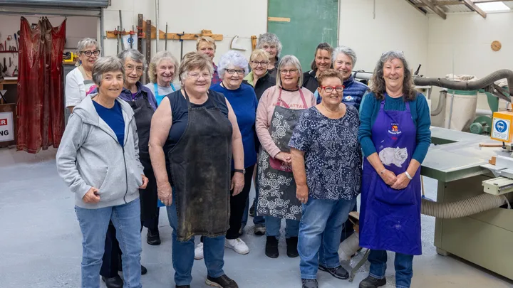 A group of women gather in a workshop, some wearing aprons, smiling and standing in front of woodworking tools.