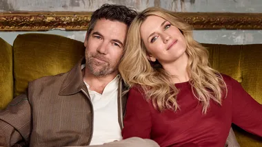 A man and woman sitting on a sofa, the woman rests her head on the man's shoulder with a relaxed expression.