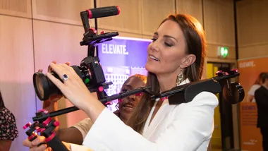 Woman in white blazer using a professional camera rig at an event, focused and smiling.