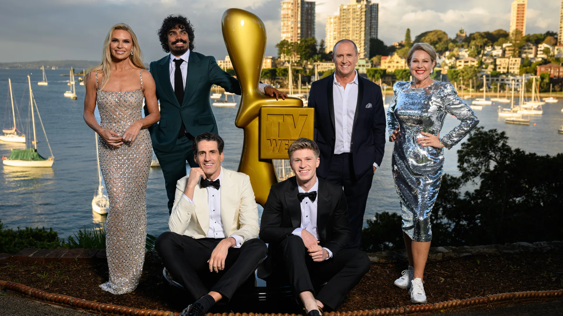 Group of six people in formal attire pose outdoors by a large TV award statue with a waterfront city skyline backdrop.