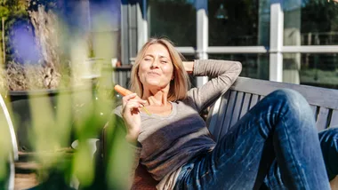 Woman relaxing on a bench outdoors, smiling and holding a carrot stick, with plants in the foreground.