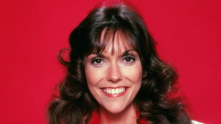 A person with long, wavy brown hair smiling against a bright red background.
