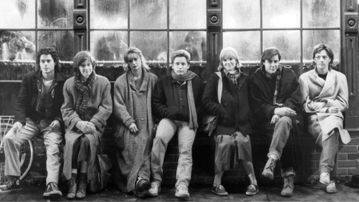 A group of seven young adults sit on a bench in front of frosted windows, dressed in winter clothing from the 1980s.
