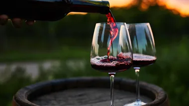 Two glasses of red wine being poured outdoors at sunset, with wine flowing from a bottle.