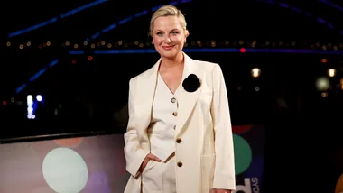 A woman in a white suit with a black flower brooch, smiling at a nighttime event with lights in the background.