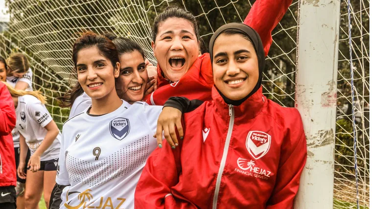 Group of women soccer players from Melbourne Victory smiling at the camera near a goalpost.