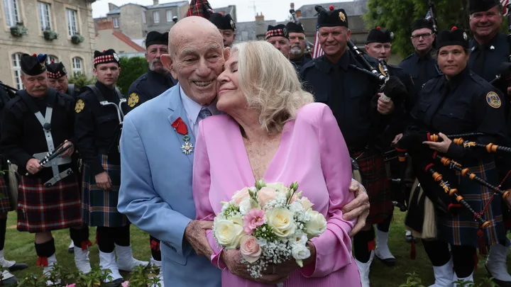 Elderly couple smiling and embracing, surrounded by bagpipers in traditional attire at a wedding ceremony.