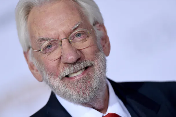 Elderly man with white hair and beard smiling, wearing glasses and a suit with a red tie.