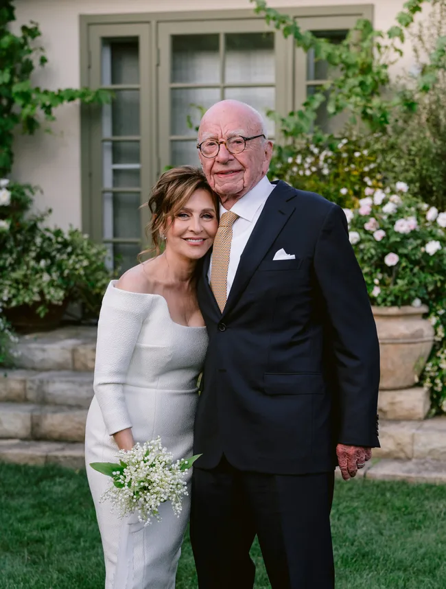 A couple poses outdoors, a woman in a white dress holding flowers, leaning on a man in a suit.