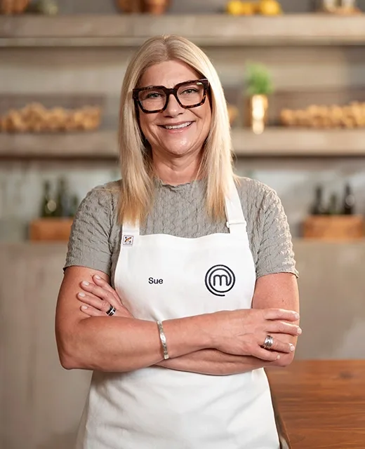 A smiling woman with glasses, wearing a "Sue" MasterChef apron, stands with folded arms in a kitchen setting.