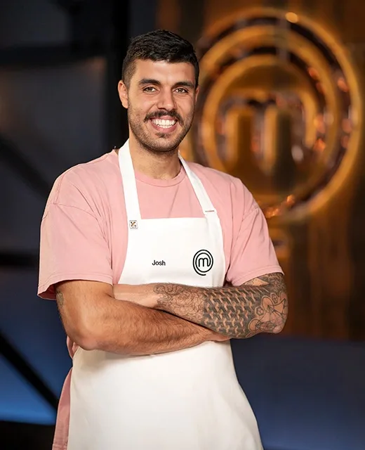 Contestant Josh smiling, in a pink shirt and white apron with MasterChef logo, standing in front of a blurred logo backdrop.