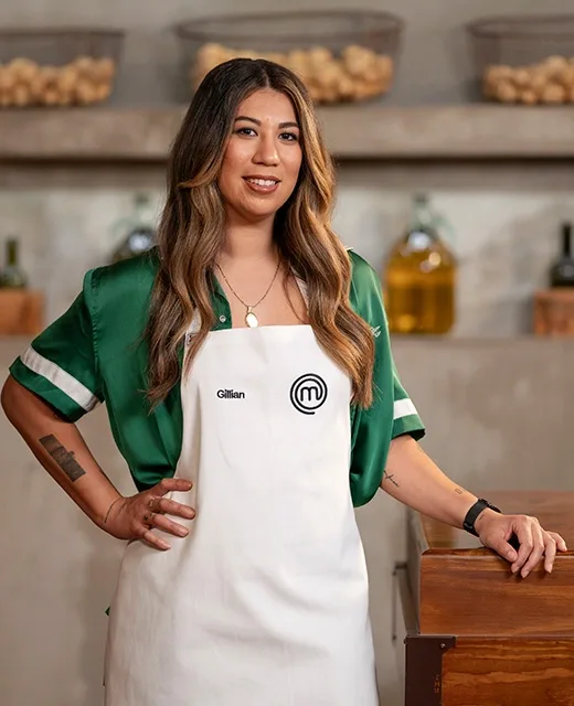 Gillian from MasterChef Australia in apron, standing in a kitchen with ingredients in the background.