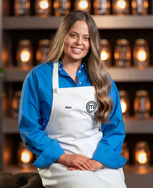 Alex from MasterChef Australia, wearing a white apron and blue shirt, smiles while sitting in front of a warmly lit shelf.