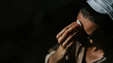 Person cleans forehead with cotton pad, wearing a headband, in dim lighting; a self-care or skincare routine.