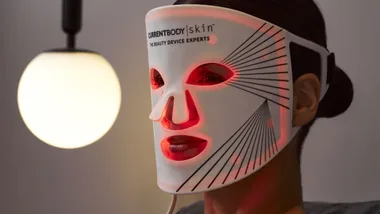Person wearing a red LED light therapy mask with "CurrentBody Skin" logo under a spherical lamp.