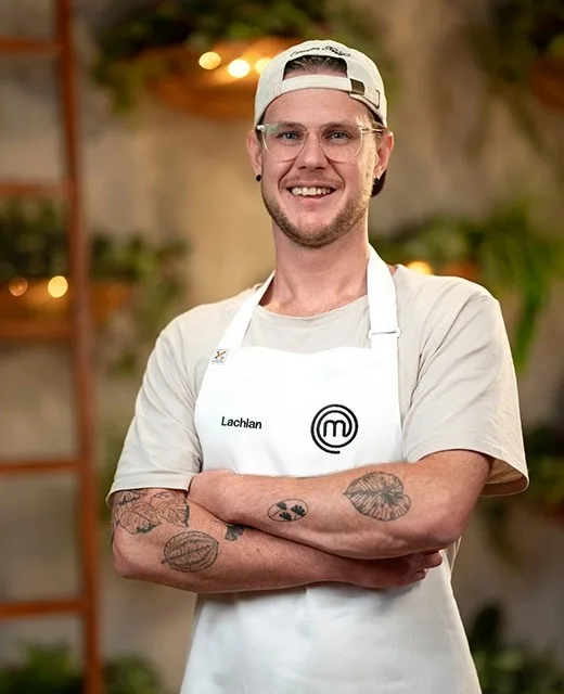 Man in a chef apron with "Lachlan" badge, smiling with arms crossed, standing in a kitchen setting.