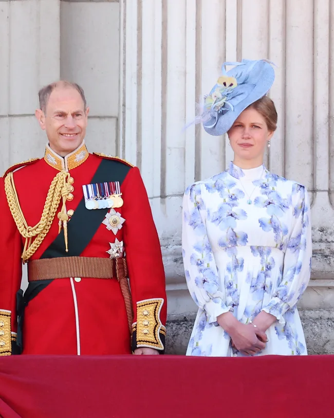 A man in a red military uniform stands next to a woman in a floral dress and blue hat, both in front of a stone column.